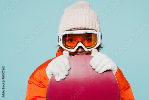 Close up young fun woman wear warm padded windbreaker jacket hat ski goggles mask hold snowboard looking camera travel rest spend weekend winter season in mountains isolated on plain blue background