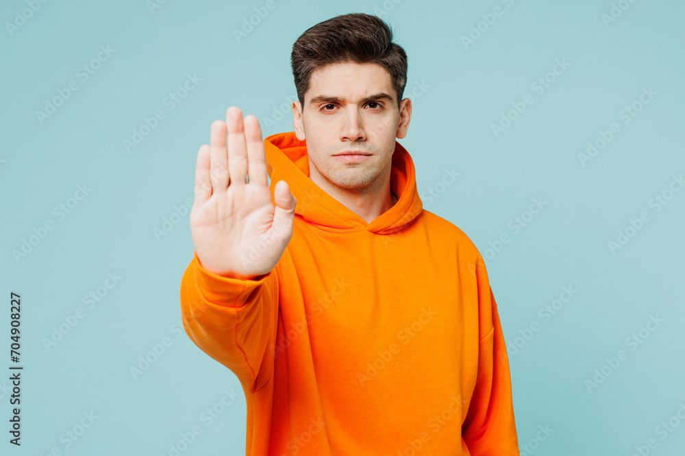 Young strict serious man he wear orange hoody casual clothes showing stop gesture with palm looking camera isolated on plain pastel light blue cyan color background studio portrait. Lifestyle concept Young strict serious man he wear orange hoody casual clothes showing stop gesture with palm looking camera isolated on plain pastel light blue cyan color background studio portrait. Lifestyle concept