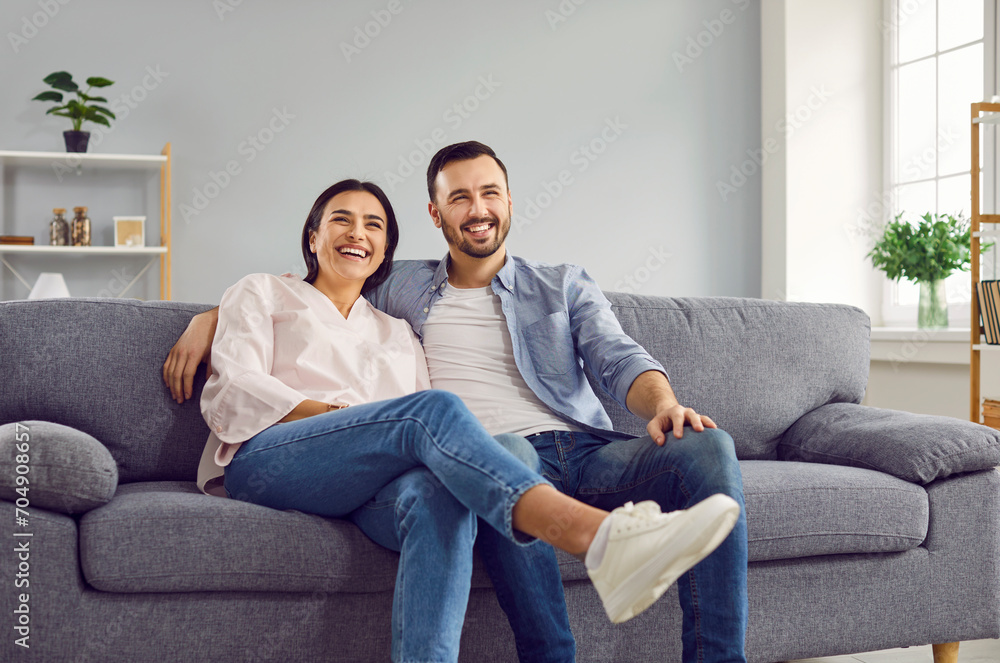 Portrait of young happy married couple sitting on the sofa in the living room and laughing. Smiling family man and woman on the couch hugging, loving wife and husband. Harmonic relationships concept. Portrait of young happy married couple sitting on the sofa in the living room and laughing. Smiling family man and woman on the couch hugging, loving wife and husband. Harmonic relationships concept.