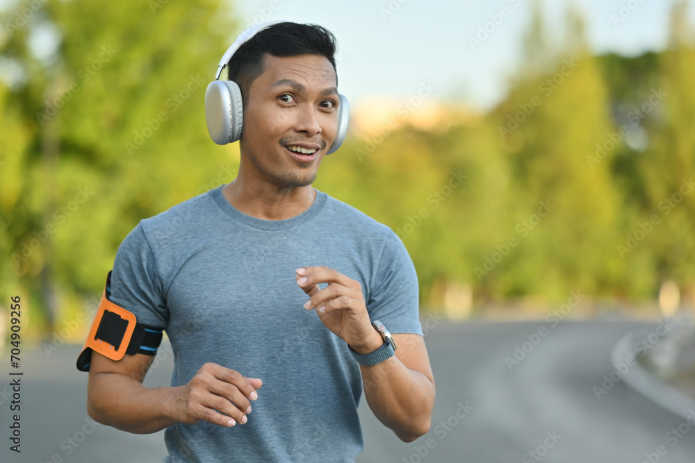Fototapeta premium Happy millennial man listening to music in headphones, jogging in the park on a sunny day