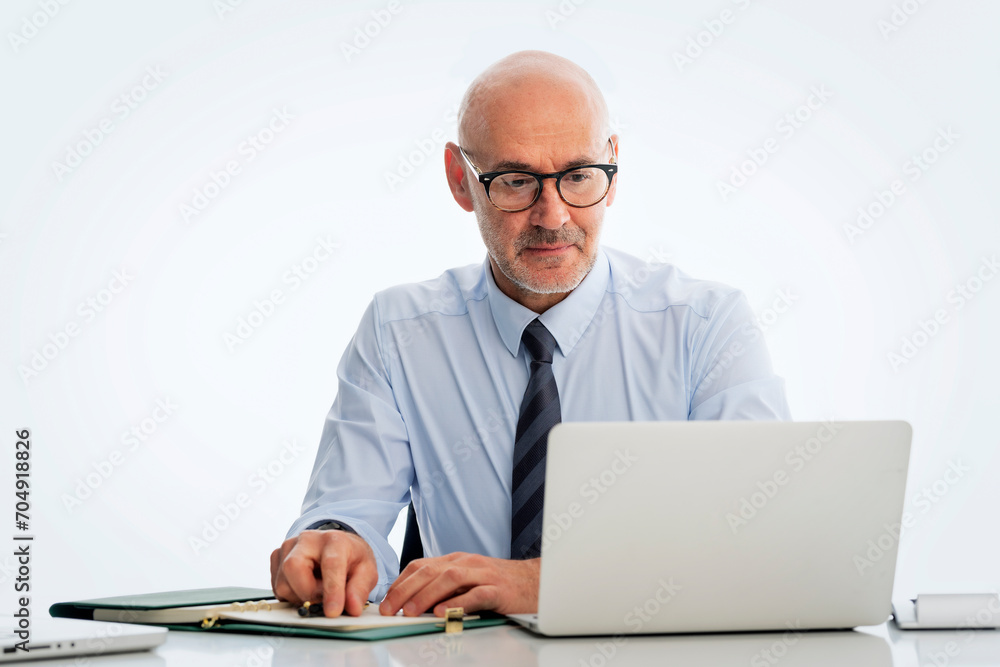 Mid aged businessman sitting at desk and using notebook for work against isolated white background Mid aged businessman sitting at desk and using notebook for work against isolated white background