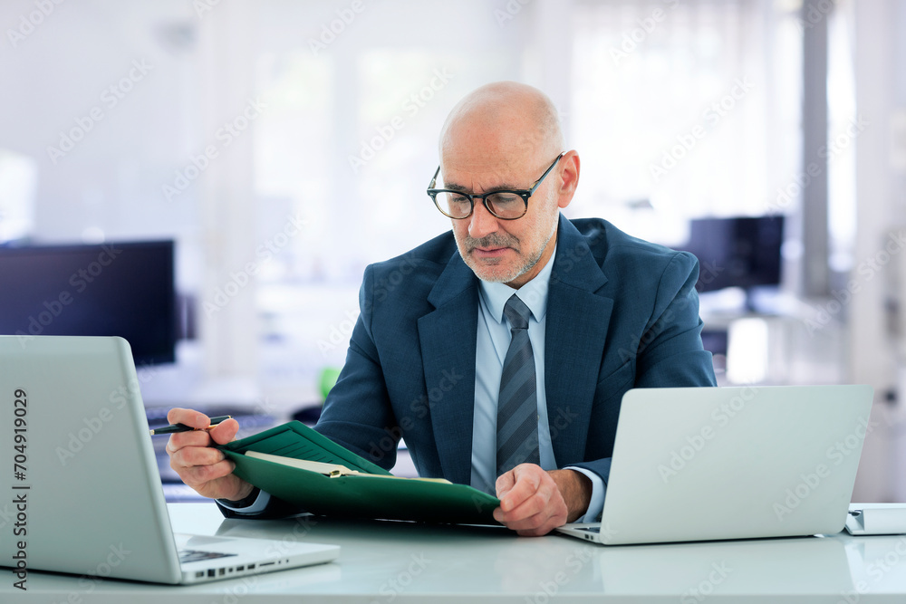 A middle-aged businessman sitting at his desk in the office and using a laptop for work A middle-aged businessman sitting at his desk in the office and using a laptop for work