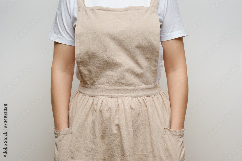 Front view of a woman in a white t-shirt and a beige linen apron against a neutral background, hands casually tucked into pockets. Front view of a woman in a white t-shirt and a beige linen apron against a neutral background, hands casually tucked into pockets.