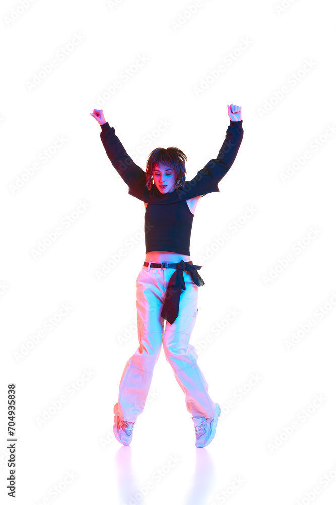 Young artistic woman in casual outfit dancing, performing hip hop isolated over white background in neon light. Concept of contemporary dance, street style, youth, hobby, action, lifestyle