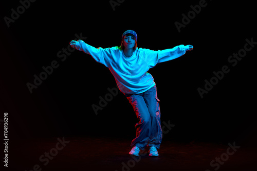 Hip hop culture. Young woman in sportive clothe dancing modern dance styles isolated over black background in neon light. Concept of contemporary dance, street style, youth, hobby, action, lifestyle