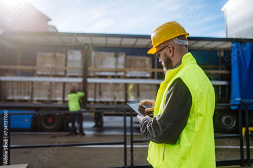 Fotografie Warehouse manager overseeing unloading of truck, holding tablet, looking at cargo details, checking delivered items, goods against order