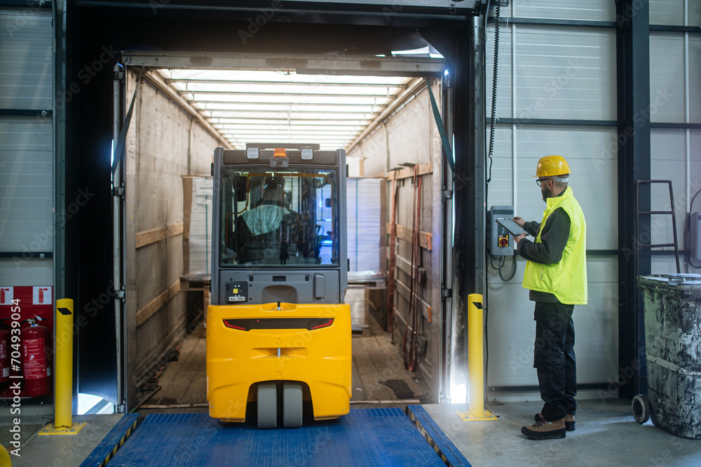 Warehouse receiver overseeing the storing of delivered items, holding tablet, looking at cargo details. Forklift carrying pallets with goods.