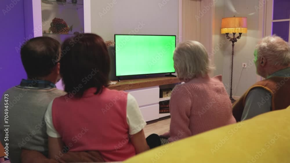 Family watching TV. Green screen. Two couples, an elderly and a middle-aged couple, are sitting on the couch at home. A woman presses a button on a TV remote control. Horizontal panning. Family watching TV. Green screen. Two couples, an elderly and a middle-aged couple, are sitting on the couch at home. A woman presses a button on a TV remote control. Horizontal panning.