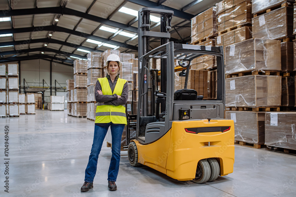 Female forklift driver standing by forklift. Warehouse worker preparing products for shipmennt, delivery, checking stock in warehouse.