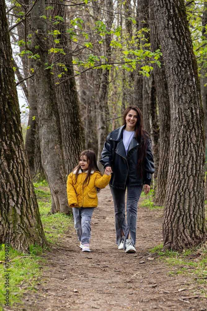 Fototapeta premium Young mother walking with her little daughter in the green park. Family holiday in garden. Portrait mom with child together on nature. Mum, little daughter outdoors. Happy Mothers Day.