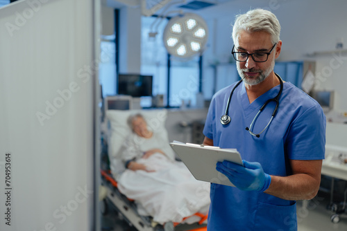 Portrait of handsome male doctor, patient in hospital bed behind. ER doctor examining senior patient, reading her medical test, lab results in clipboard. photo