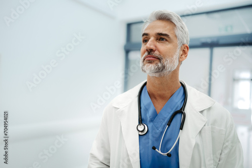 Portrait of confident mature doctor standing in Hospital corridor. Handsome doctor with gray hair wearing white coat, stethoscope around neck standing in modern private clinic, looking at camera.