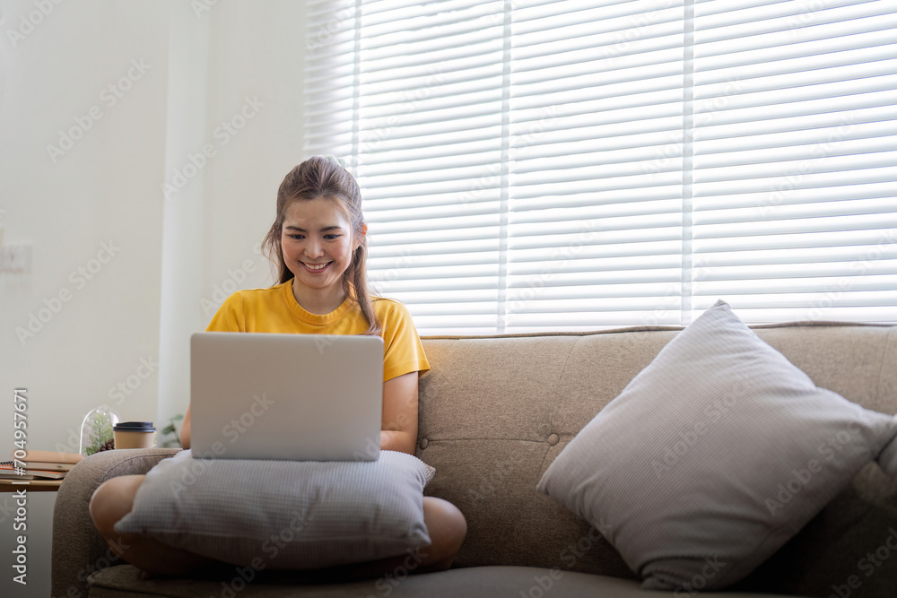 Young woman sitting on the couch and working on project, watching movie on laptop rest and happy chatting with friend in social network at home