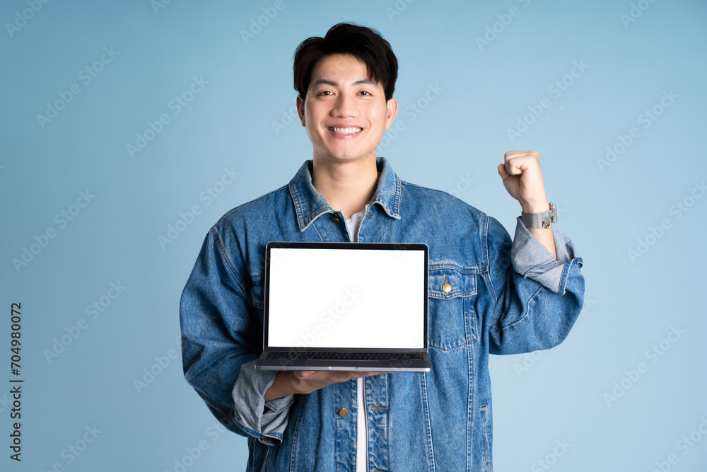 Portrait of an Asian guy wearing a jacket and using a laptop posing on a blue background Portrait of an Asian guy wearing a jacket and using a laptop posing on a blue background