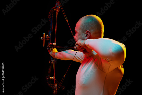 Back view image of man, archery at hele aiming with archery bow on target against black studio background in neon light. Concept of professional sport and hobby, competition, action, game photo