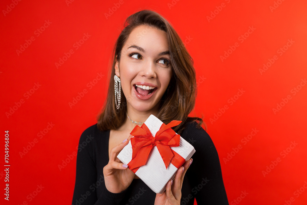 Fototapeta premium Valentine's day. A young woman gives a gift in a white box. Studio portrait on a red background