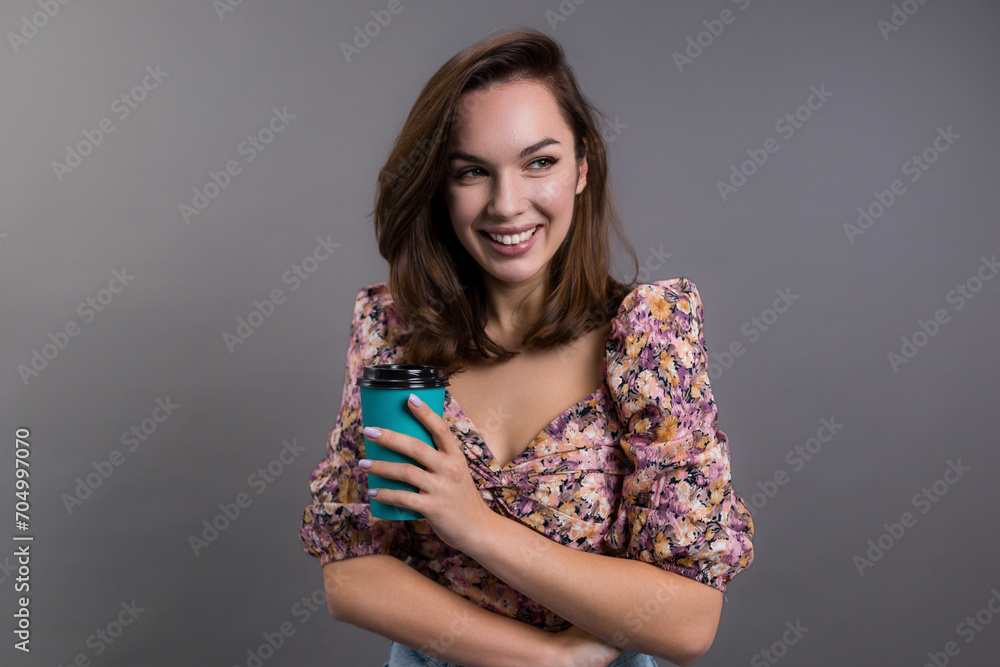 An attractive young woman in stylish clothes holds a paper eco coffee cup on a gray background. The model smiles and looks away An attractive young woman in stylish clothes holds a paper eco coffee cup on a gray background. The model smiles and looks away