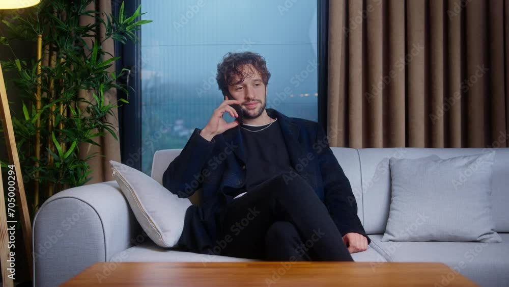 Young curly hair businessman speaking using mobile phone, sitting on sofa in living room at home. Male relaxing at home, communicating with family and friends Young curly hair businessman speaking using mobile phone, sitting on sofa in living room at home. Male relaxing at home, communicating with family and friends