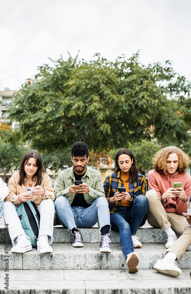 Fototapeta premium Vertical photo. Millennial group of diverse young people using mobile phone outside. Connected student friends on smartphone devices sitting in city street. Technology lifestyle concept