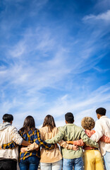 Vertical photo. Rear view of young group of happy multiracial friends hugging each other standing in a row looking blue sky