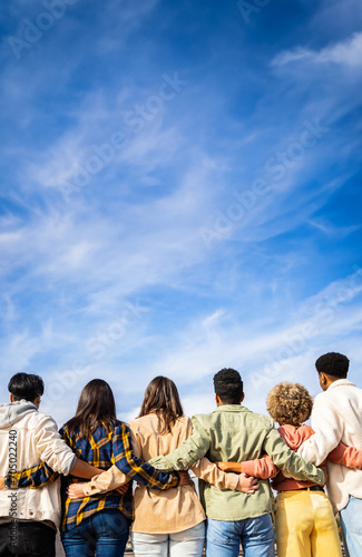 Vertical photo. Rear view of young group of happy multiracial friends hugging each other standing in a row looking blue sky