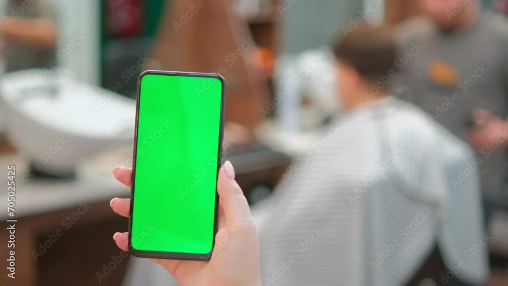 Barbershop. Back view of brunette holding chroma key green screen smartphone watching content. Gadgets and contemporary people concept. 4K Barbershop. Back view of brunette holding chroma key green screen smartphone watching content. Gadgets and contemporary people concept. 4K