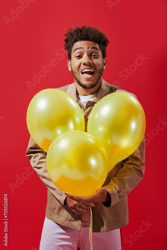 positive african american man in beige jacket holding balloons and laughing on red background © LIGHTFIELD STUDIOS