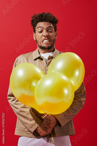 funny african american man in beige jacket looking at balloons and grimacing on red background © LIGHTFIELD STUDIOS