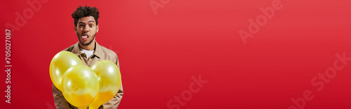 banner, african american man in beige jacket holding balloons and sticking tongue on red background © LIGHTFIELD STUDIOS