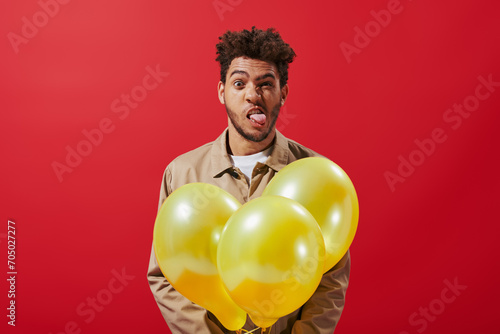 funny african american man in beige jacket holding balloons and sticking tongue on red background © LIGHTFIELD STUDIOS