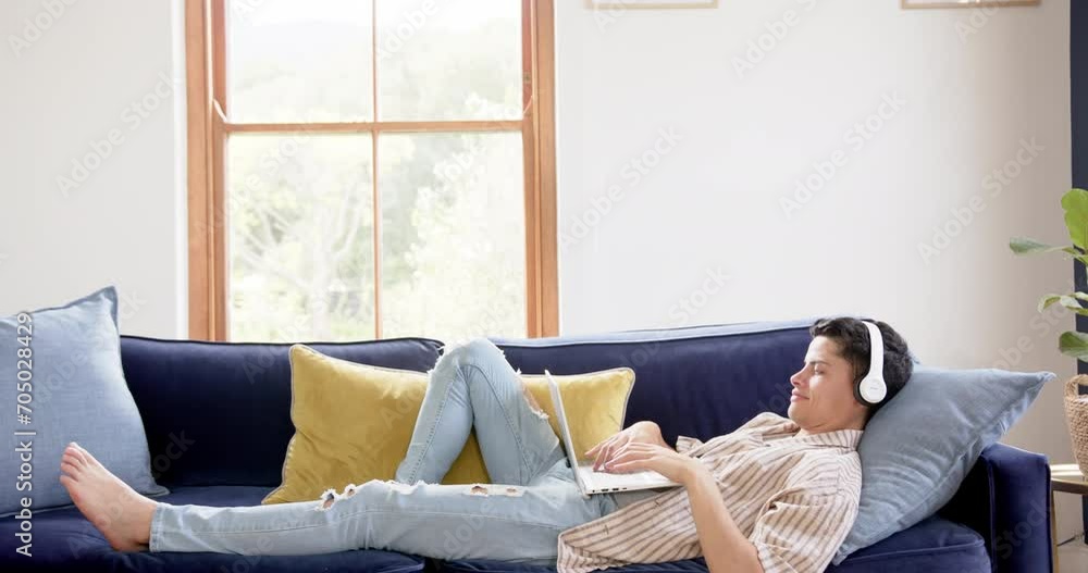 Happy biracial man wearing headphones lying on couch using laptop at home, copy space, slow motion Happy biracial man wearing headphones lying on couch using laptop at home, copy space, slow motion