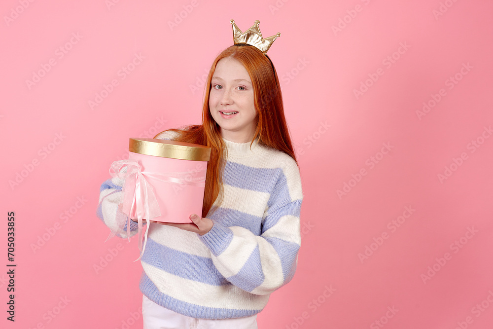 Smiling redhead teenage girl wearing crown and sweater holding present box on pink background in studio. Smiling redhead teenage girl wearing crown and sweater holding present box on pink background in studio.