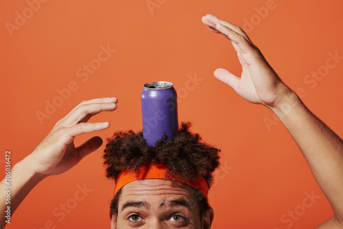 purple soda can on head of curly african american man with headband on orange background, gesture