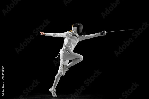 Confidence and strength. Female athlete in fencing gear showcasing her impeccable form, sword poised for action against a black studio background. Concept of active and healthy lifestyle, championship