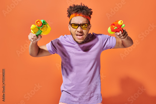 tensed african american fella in sunglasses holding water guns on orange background  summer fun