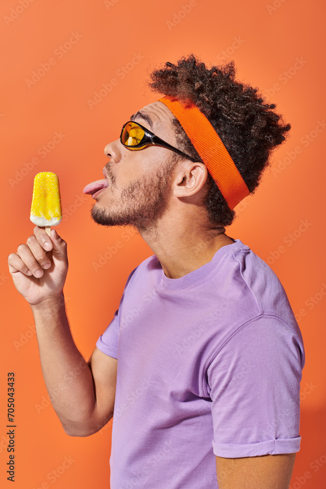 african american man in sunglasses and headband licking frozen ice cream on orange backdrop, tongue