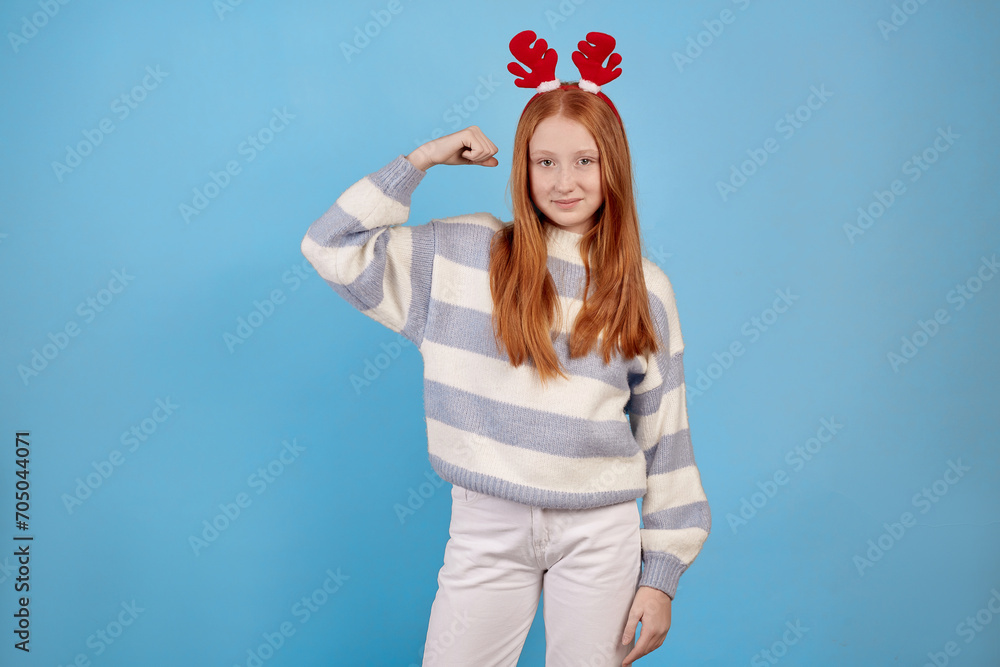 Smiling redhead teenage girl with decorative deer horns raises arms and shows biceps on blue background in studio. Smiling redhead teenage girl with decorative deer horns raises arms and shows biceps on blue background in studio.