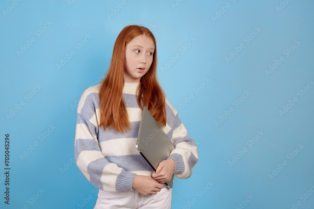 Confident redhead teenage girl holding laptop on blue background in studio.