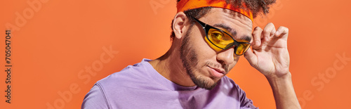 banner, young african american man adjusting sunglasses and looking at camera on orange background photo