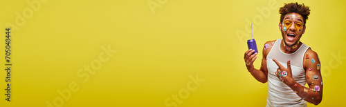 excited african american man with stickers on face pointing at soda can on yellow background