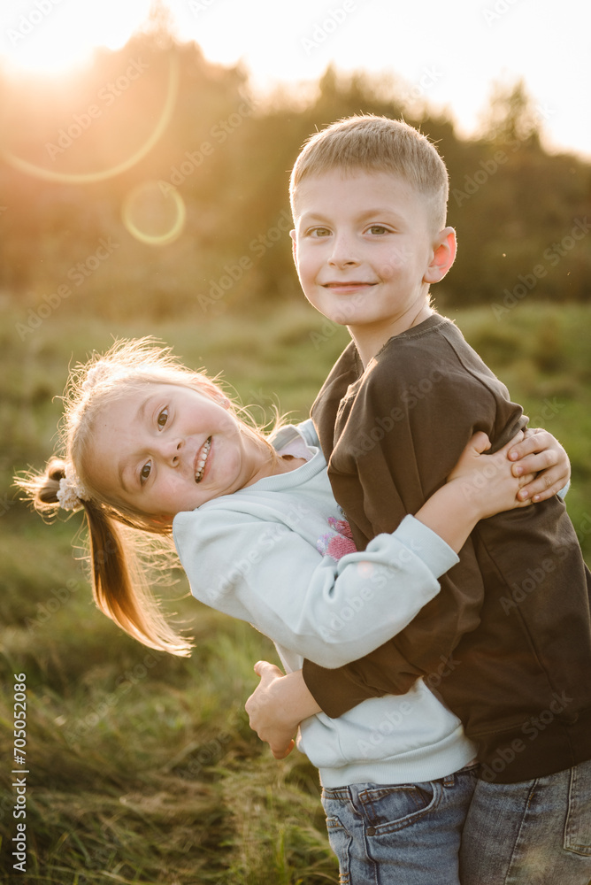 Fototapeta premium Daughter, son hugging in green grass in field at sunset. Happy children walking spending time together in nature. Concept family holiday outdoors. Kids playing in mountains on autumn day. Closeup