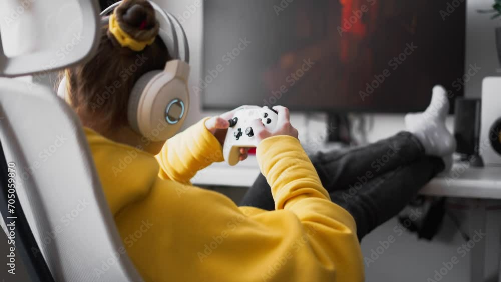 Teenage girl with headset is sitting in a comfortable computer chair, holding a white gamepad in her hands and playing a game on a PC or console. Live Hand Held shooting. Teenage girl with headset is sitting in a comfortable computer chair, holding a white gamepad in her hands and playing a game on a PC or console. Live Hand Held shooting.