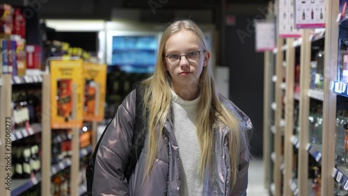 Young girl standing in between alcoholic drinks shelves. Blond hair girl watching into the camera. Global alcohol drinking problem concept.