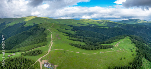 Aerial view above the vast mountain sides and mountaintops of Capatanii Mountains. Green pastures are crossed by numerous dirt roads. Wild spruce forests grow between the crests, along the valley.  photo