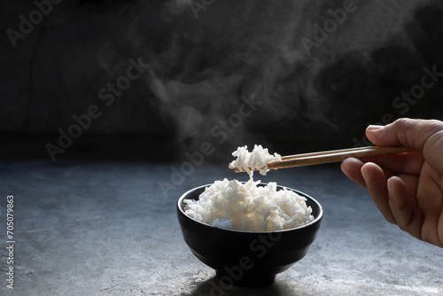The man hand of using black chopsticks holding hot jasmine rice with smoke and steam in black bowl over dark background.hot food concept photo
