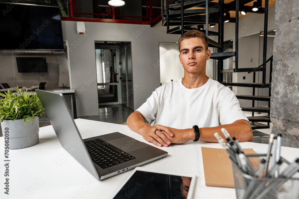 Casual young man in white t-shirt sitting at white desk looking at laptop screen at modern office