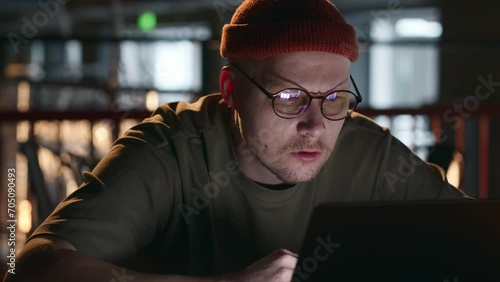 A handsome man with glasses is a millennial hacker sitting at a laptop in a dark room and swearing at the screen. A computer hacker or an Internet scammer does not understand what is happening. 4k photo