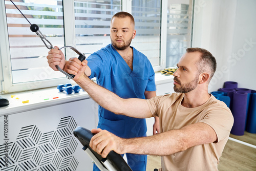 young rehabilitologist in blue uniform helping patient during recovery training in kinesio center photo