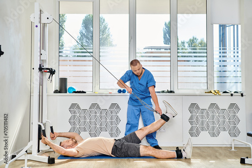 skilled rehabilitologist assisting man working out on exercise machine in gym of kinesio center