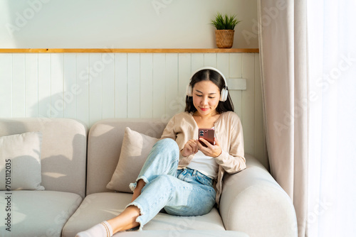 Happy young Asian woman with mobile phone listening music in headphones and relaxing on safa at home